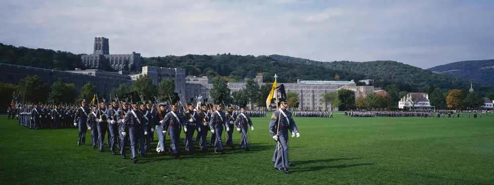 Soldiers in dress uniforms march in formation across a grassy parade ground during an army graduation ceremony.