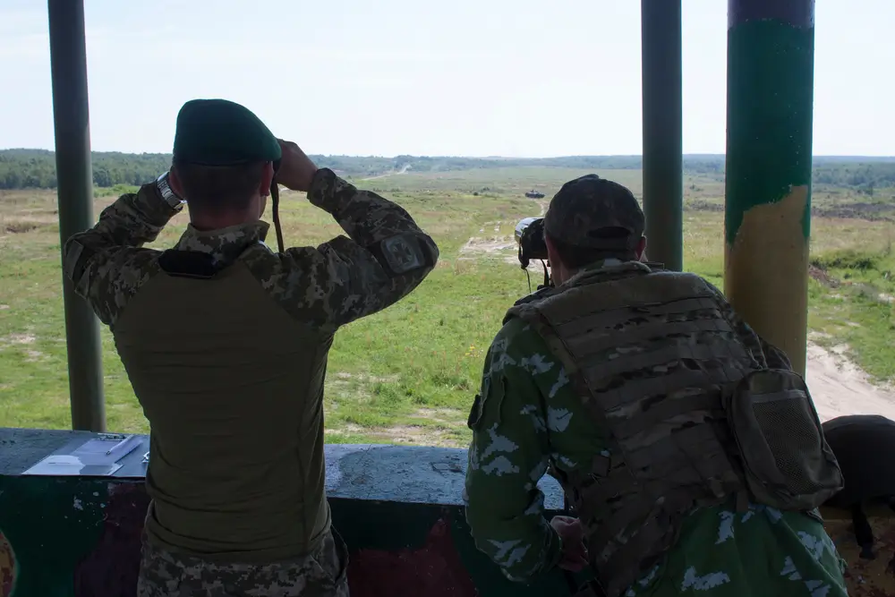 Army reservists scan an open military field with binoculars and a spotting scope during a field exercise.