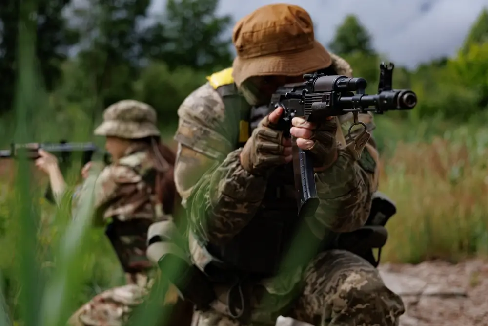 Infantry soldiers in camouflage gear aiming rifles during tactical field training at a military camp.