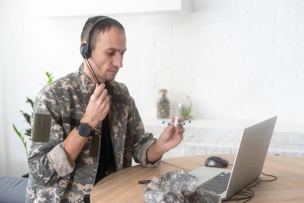 Soldier in uniform wearing a headset takes a military aptitude test on a laptop while holding a model airplane.