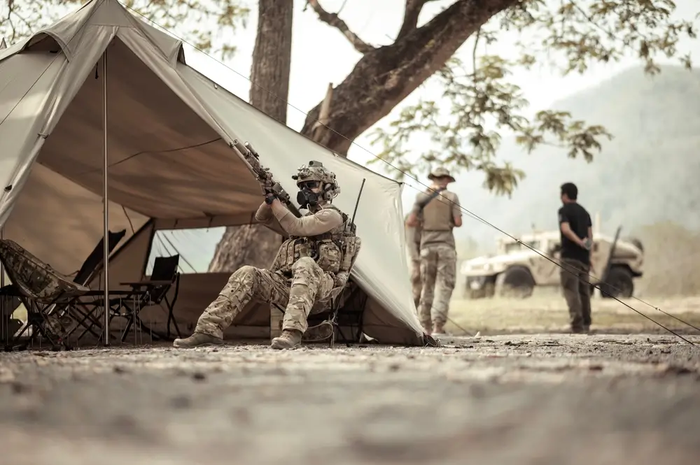 Soldier in tactical gear at a military camp army base, sitting by a tent with a vehicle and personnel in the background.