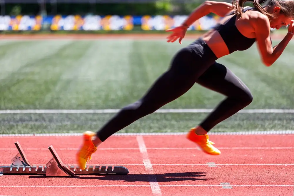 Athlete in yellow running shoes sprinting from starting blocks on a track, often used for fitness tests over a gym floor.