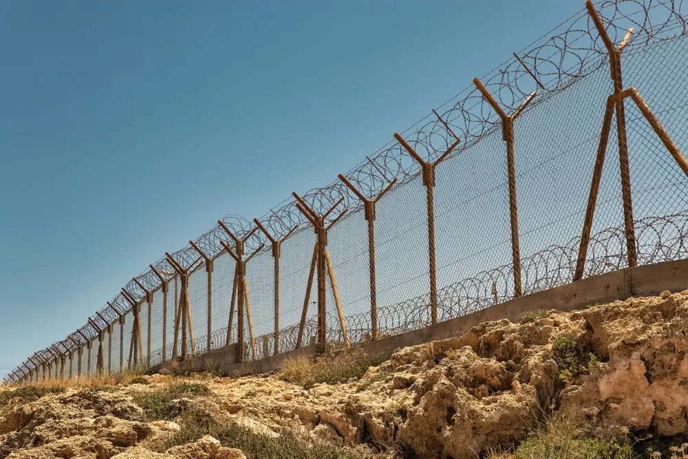 Secure perimeter of a military base featuring a tall chain-link fence topped with razor wire on rocky ground.