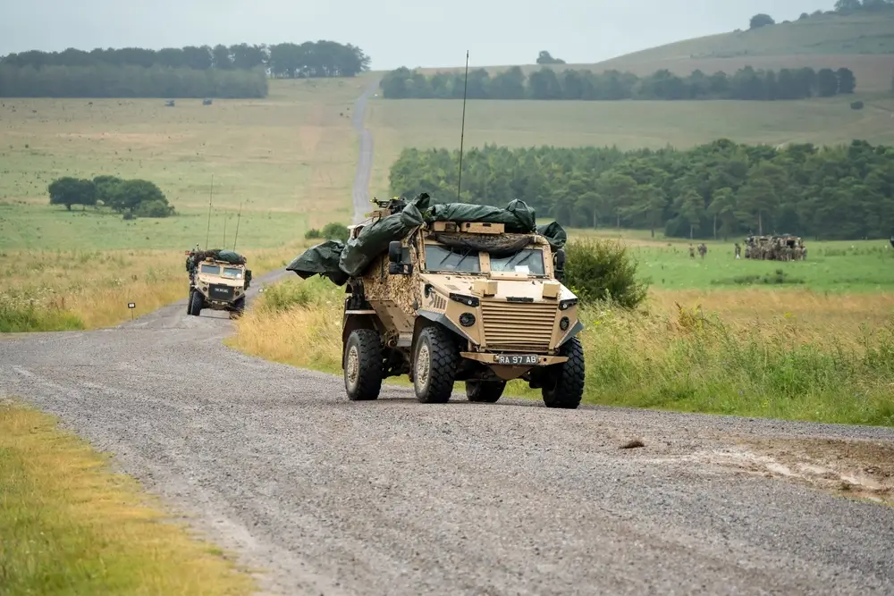 Military light armoured vehicles driving on a gravel road in a rugged rural army training area.