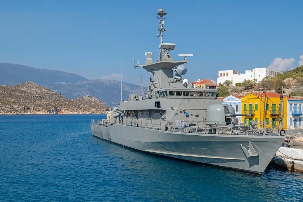Modern naval warship docked at a navy base port with colorful houses and mountains in the background.