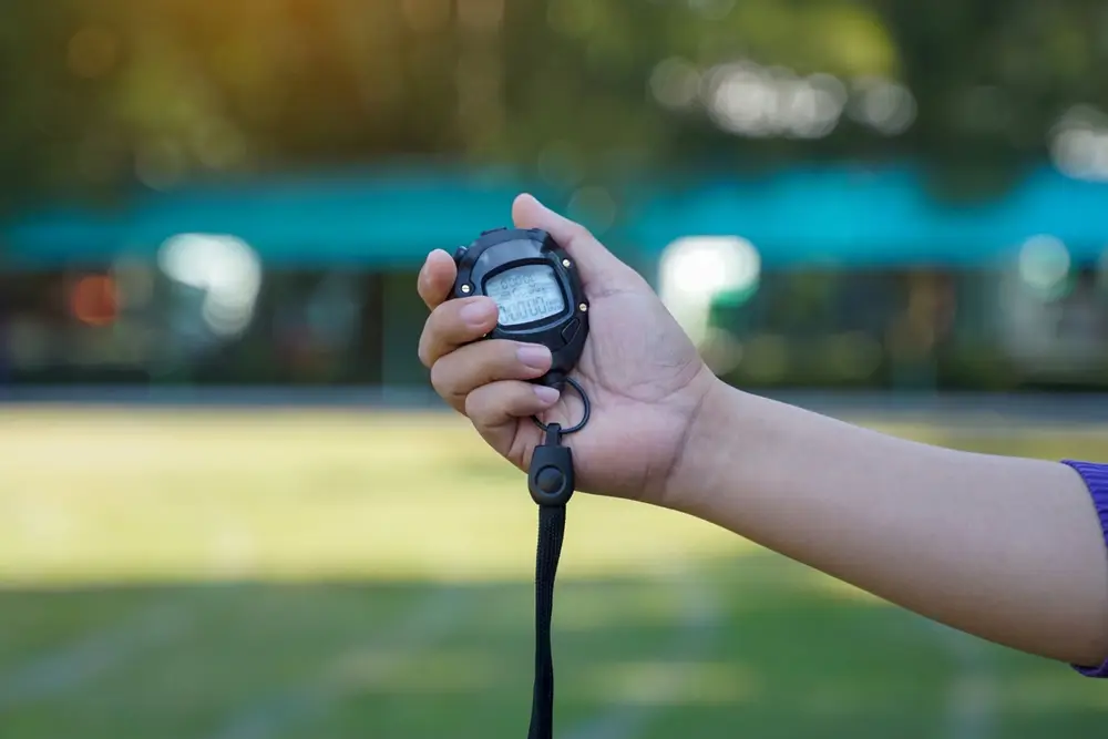 Hand holding a digital stopwatch for personal trainer fitness coaching during a timed outdoor shuttle run fitness test.