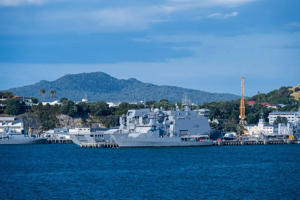 Royal New Zealand Navy ships docked at a government military port facility with coastal hills in the background.