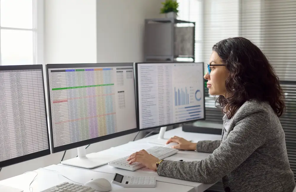 Focused professional analyzing numerical reasoning tables and business charts on multiple screens for data analytics.