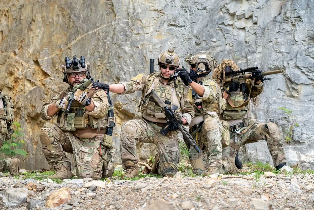 Soldiers in tactical gear perform a military training drill, kneeling with rifles during a base field exercise.