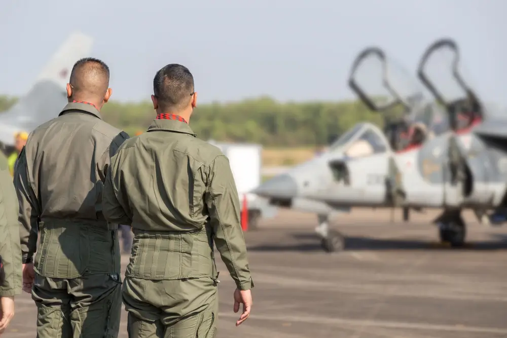 Military pilots in flight suits look toward a jet aircraft during a briefing before a training flight.