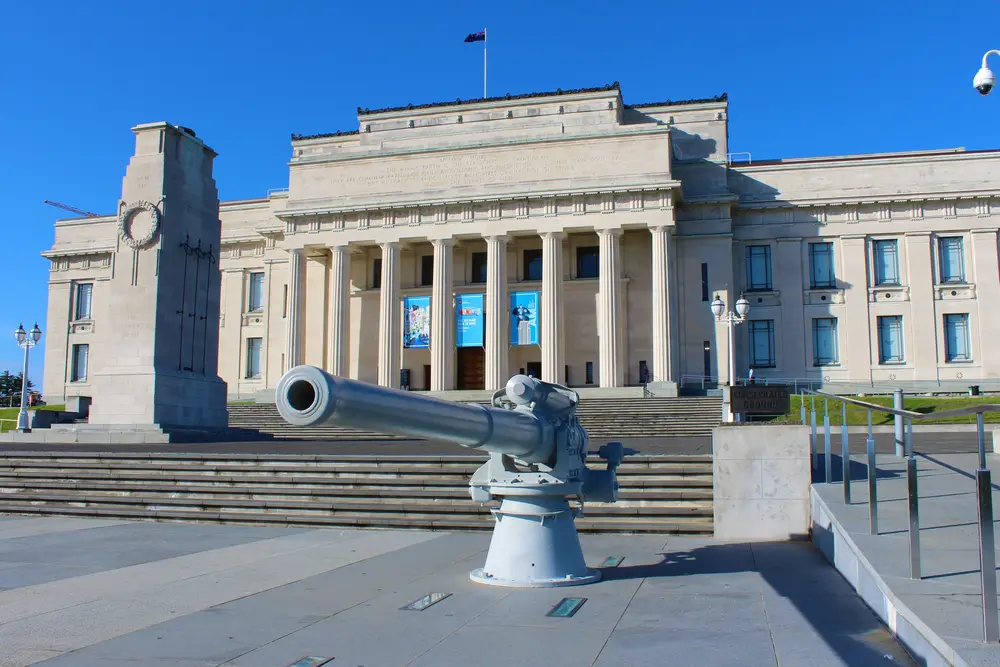 Artillery gun in front of a New Zealand military base building with stone columns and the national flag flying.
