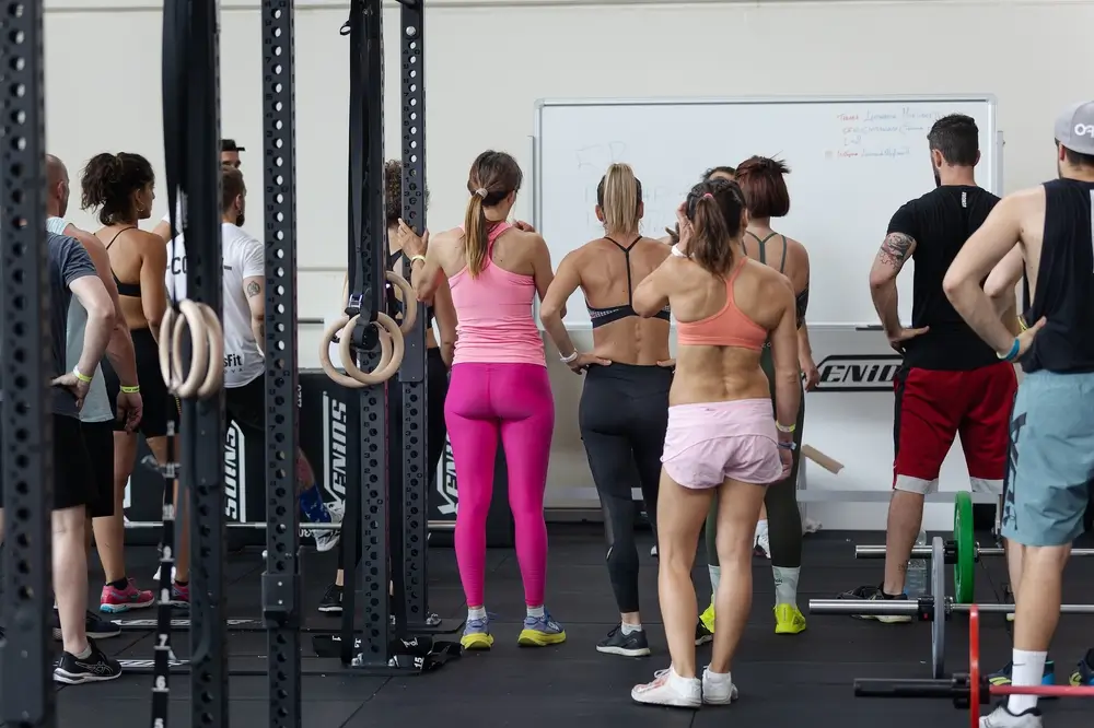 Fitness recruits in sports gear gather around a whiteboard during a group physical training session in a gym.