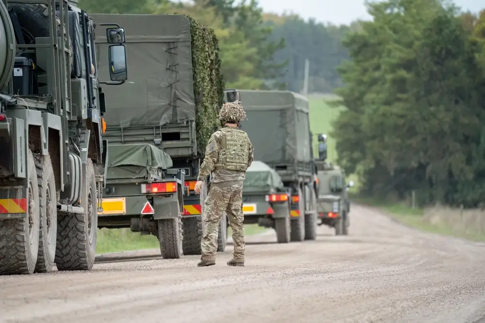 Soldier in camouflage stands next to a line of military army logistics trucks on a dirt road at a training camp.