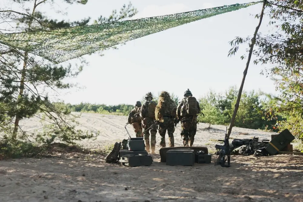 Soldiers in full gear walk across a sandy field under a camouflage net at a military training camp.