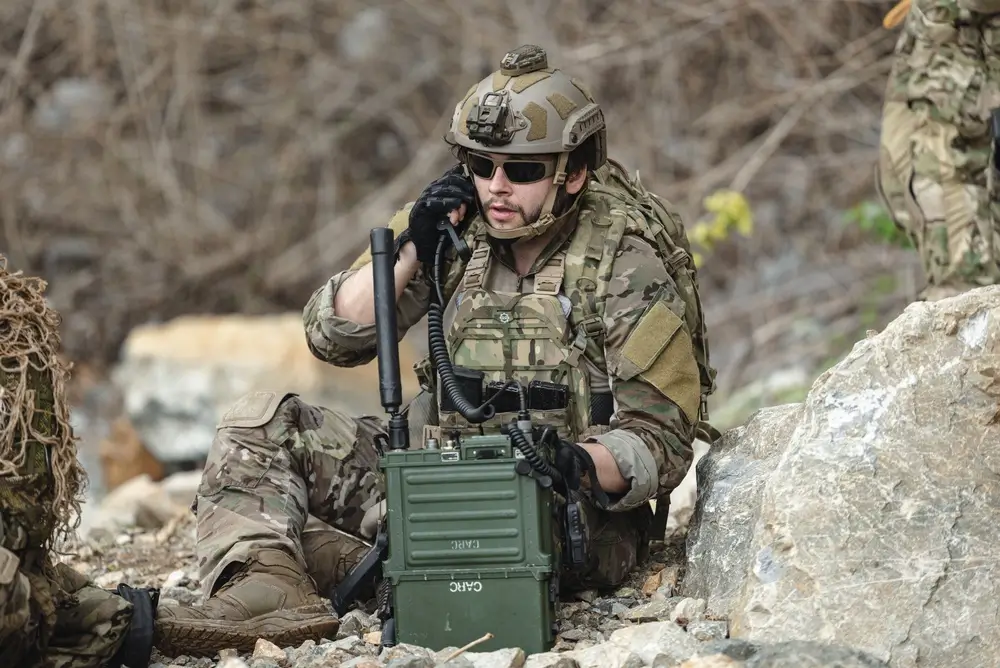 Soldier using field radio equipment and military communication devices during training in a rocky landscape.