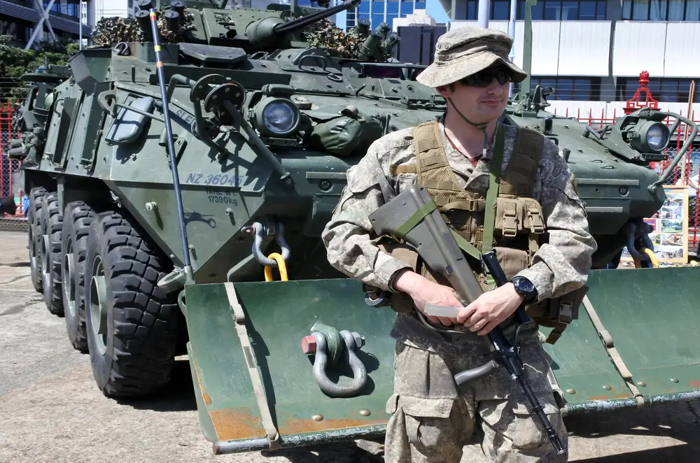 New Zealand Army soldier in NZMTP combat uniform and jungle hat, holding a rifle in front of an armored vehicle.
